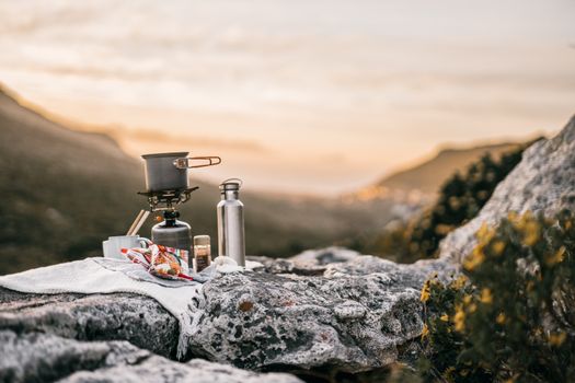 Équipement de camping sur une roche avec un réchaud, une bouteille d'eau et des collations au coucher du soleil.