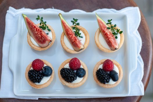 Assiette blanche avec six tartelettes garnies de crème, fruits rouges et figues, sur un napperon.