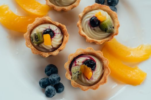 Bouchées sucrées en tartelette garnies de crème, fruits et myrtilles sur une assiette blanche.