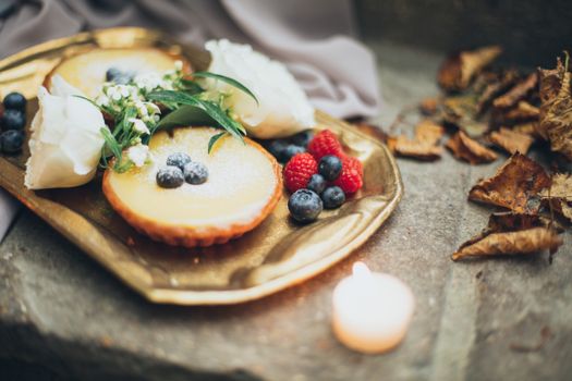 Plateau doré avec tarte aux myrtilles et poires, décoré de fruits rouges et fleurs, ambiance chaleureuse avec une bougie.