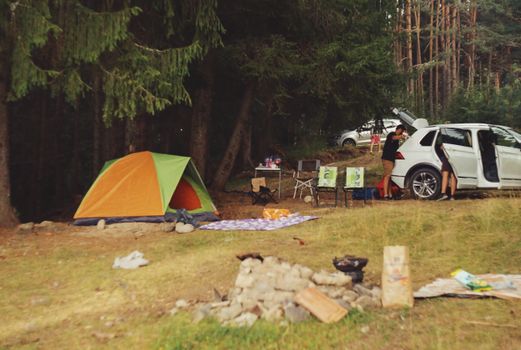 Camping en pleine nature avec une tente orange et verte, une voiture, des chaises et un feu de camp.