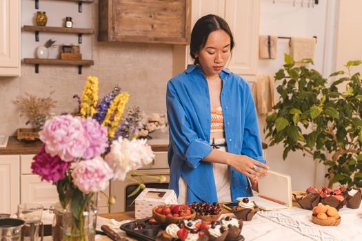 Femme en chemise bleue préparant des bouchées sur une table décorée avec des fleurs et des desserts variés.