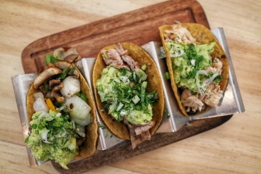Trois taquitos garnis de poulet, pommes de terre, guacamole et herbes fraîches sur une planche en bois.